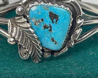 May include: A silver cuff bracelet featuring a vibrant turquoise stone at its center. The stone is a striking shade of blue with dark brown inclusions. The bracelet has intricate metalwork, including a leaf design and decorative edging.