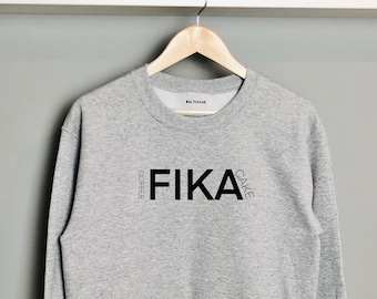May include: Gray sweatshirt with the word "Fika" printed in black, with "Coffee" and "Cake" written vertically on either side of the word.