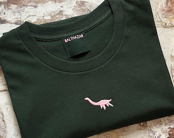 Könnte beinhalten: Ein dunkelgrünes T-Shirt mit einem rosa gestickten Dinosaurier auf der Vorderseite. Das T-Shirt hat einen Rundhalsausschnitt und das Label "BALTHAZAR" ist am Kragen sichtbar.