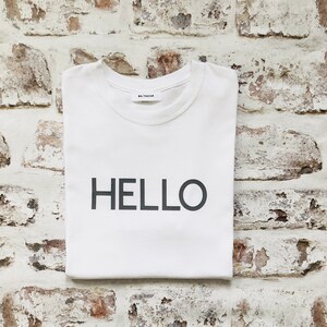 Puede incluir: Camiseta blanca con la palabra "HELLO" impresa en negro en la parte delantera. La camiseta tiene una pequeña etiqueta con la palabra "BALTHAZAR" en el lado izquierdo del pecho.