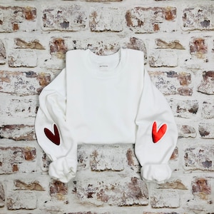 Peut inclure: Sweat-shirt blanc avec des patchs en forme de cœur rouge sur les coudes. Le sweat-shirt a un col rond et des manches longues.