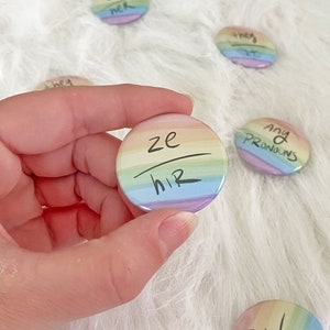 May include: A collection of rainbow-colored pronoun buttons. One button is held in a hand, displaying "ze/hir" in black ink. Other buttons show pronouns like "she/her", "they/them", "he/him", and "any pronouns".