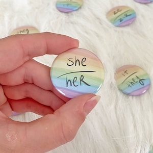 May include: A collection of round, rainbow-colored pronoun buttons. One button is held in a hand, displaying "she/her" in black script. Other buttons show "they/them", "he/him", and "ze/zir". The buttons are on a white, fluffy surface.
