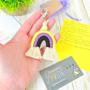 May include: A handmade non-binary pride flag keychain. The rainbow-shaped macrame keychain features yellow, purple, and black colors, with a silver clasp and wooden beads. The keychain has a tassel fringe at the bottom.