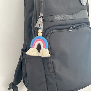 May include: A colorful rainbow macrame keychain with blue, pink, and white arches and cream-colored fringe. The keychain is attached to a silver zipper pull and wooden beads. The keychain is hanging on a black backpack.