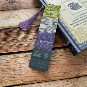 May include: A colorful bookmark featuring a bookshelf design with a purple tassel. The bookmark has a series of shelves filled with books in various shades of purple, gray, and green. A globe and decorative vase are also depicted.