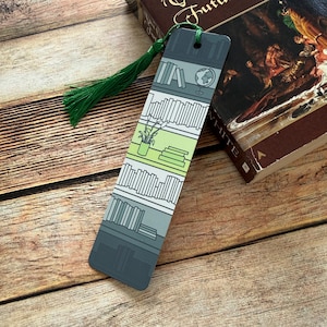 May include: A decorative bookmark featuring a bookshelf design with a green tassel. The bookmark has a gray and green color scheme, with illustrations of books, a globe, and a potted plant. The bookmark is resting on a wooden surface next to a book.
