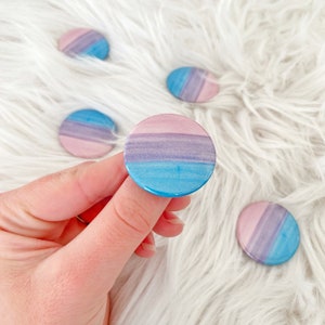 May include: A collection of round, colorful buttons with a watercolor design. The buttons feature a gradient of blue, purple, and pink hues. One button is held by a hand, while others are scattered on a white, fluffy surface.