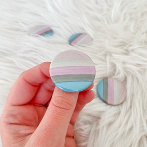 May include: A collection of round, colorful buttons with a watercolor design. The buttons feature horizontal stripes in shades of blue, gray, pink, and white. One button is held between fingers, with others scattered in the background.