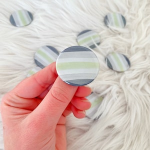 May include: A collection of round buttons with a striped design in shades of gray, white, and light green. The buttons are held in a hand and are scattered on a fluffy white surface. The buttons have a glossy finish.