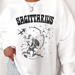 May include: White sweatshirt featuring a black Sagittarius design. The graphic includes the word "SAGITTARIUS", an archer figure, stars, and the Sagittarius constellation. A casual, comfortable top.