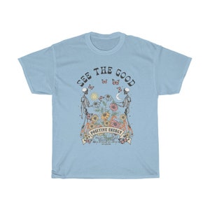 May include: Light blue t-shirt with a graphic featuring two skeletons holding hands, surrounded by flowers and butterflies. The text "SEE THE GOOD" and "POSITIVE ENERGY" are displayed above and below the central image.