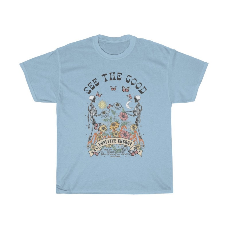 May include: Light blue t-shirt with a graphic featuring two skeletons holding hands, surrounded by flowers and butterflies. The text "SEE THE GOOD" and "POSITIVE ENERGY" are displayed above and below the central image.
