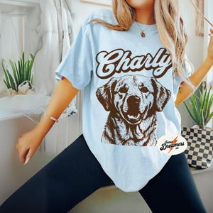May include: Light blue t-shirt with a brown graphic of a dog and the text "Charly" in a retro font. The shirt has a distressed look and is made of a soft, comfortable material.