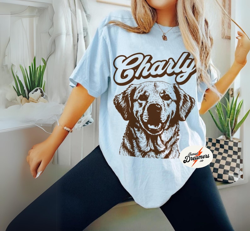 May include: Light blue t-shirt with a brown graphic of a dog and the text "Charly" in a retro font. The shirt has a distressed look and is made of a soft, comfortable material.