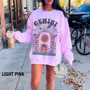Pode incluir: Uma camisola rosa claro com uma estampa gr&aacute;fica do signo do zod&iacute;aco G&eacute;meos. O gr&aacute;fico apresenta um sol, flores e duas figuras com o texto "Imaginative & Devious".