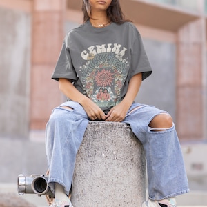 May include: A grey t-shirt with the word "GEMINI" in a retro font, featuring a colourful graphic design. The person is wearing light blue ripped jeans and white trainers. The overall style is casual and trendy.