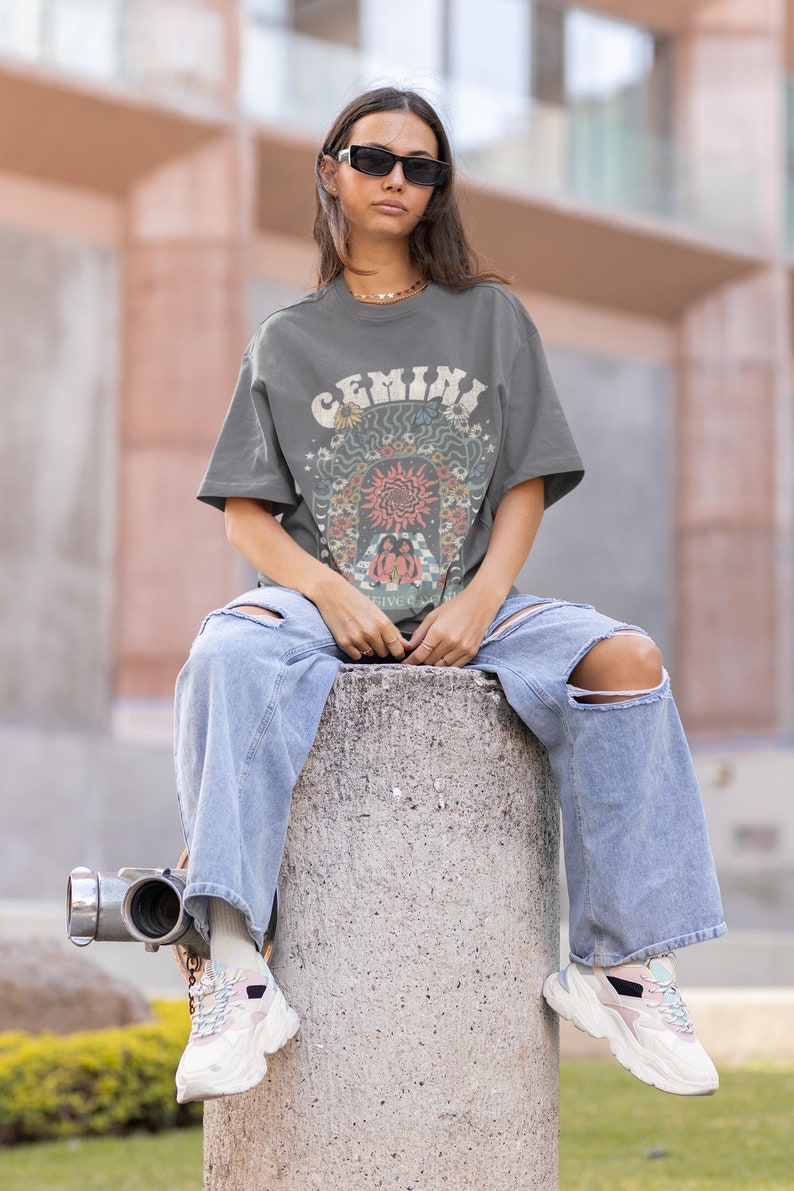 May include: A grey t-shirt with the word "GEMINI" in a retro font, featuring a colourful graphic design. The person is wearing light blue ripped jeans and white trainers. The overall style is casual and trendy.