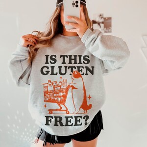 May include: A gray sweatshirt with a graphic of a cat pushing a shopping cart full of food items. The text "IS THIS GLUTEN FREE?" is printed on the sweatshirt.