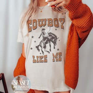 May include: Cream-colored t-shirt with the text "COWBOY LIKE ME" in brown lettering, featuring a cowboy riding a horse graphic. The shirt is paired with an orange sweater. The phone case has a pumpkin pattern.
