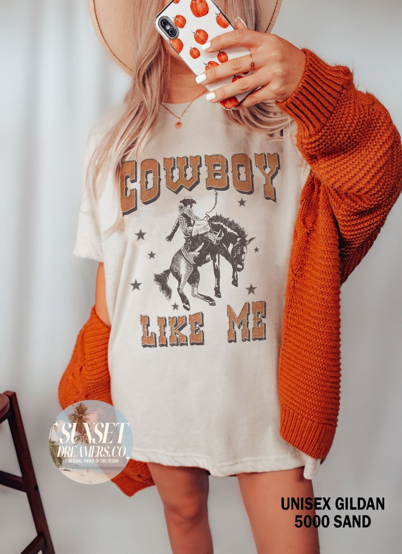 May include: Cream-colored t-shirt with the text "COWBOY LIKE ME" in brown lettering, featuring a cowboy riding a horse graphic. The shirt is paired with an orange sweater. The phone case has a pumpkin pattern.