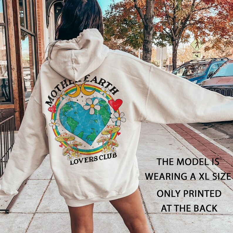 May include: A cream-colored hoodie with a graphic print on the back. The graphic features a heart-shaped earth with a rainbow, flowers, and mushrooms. The text "Mother Earth Lovers Club" is printed below the graphic.