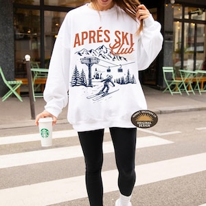 May include: White sweatshirt with a graphic of a skier on a mountain with the text "Après Ski Club" in brown and blue lettering. The graphic also includes a ski lift and trees. The sweatshirt has a brown tag with the text "Sunset Dreamers Co. Original Design".