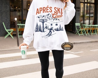Puede incluir: Sudadera blanca con un gráfico de un esquiador en una montaña con el texto "Après Ski Club" en letras marrones y azules. El gráfico también incluye un telesilla y árboles. La sudadera tiene una etiqueta marrón con el texto "Sunset Dreamers Co. Original Design".