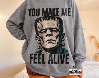 May include: A heather gray crewneck sweatshirt featuring a black and white graphic of Frankenstein's monster. The text "YOU MAKE ME" is printed above the face, and "FEEL ALIVE" is printed below in a bold, distressed black font style.