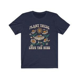 May include: A navy blue t-shirt with a retro floral design, including daisies and bees. The text "PLANT THESE SAVE THE BEES" is printed above and below the graphic in a distressed font. The shirt has short sleeves and a crew neck.