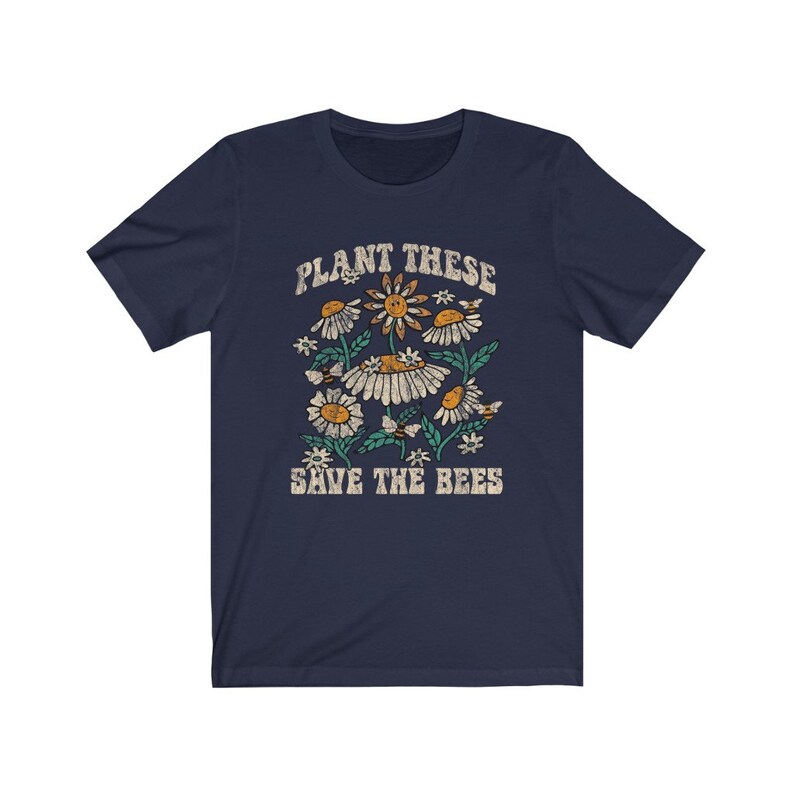May include: A navy blue t-shirt with a retro floral design, including daisies and bees. The text "PLANT THESE SAVE THE BEES" is printed above and below the graphic in a distressed font. The shirt has short sleeves and a crew neck.