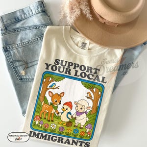May include: Cream-colored t-shirt with the text "SUPPORT YOUR LOCAL IMMIGRANTS" in a blue-outlined box. The graphic features a deer, duck, and sheep holding hands. Light blue jeans and a tan hat are also in the image.