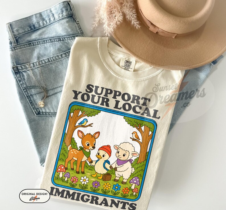 May include: Cream-colored t-shirt with the text "SUPPORT YOUR LOCAL IMMIGRANTS" in a blue-outlined box. The graphic features a deer, duck, and sheep holding hands. Light blue jeans and a tan hat are also in the image.