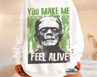 May include: A white crewneck sweatshirt featuring a distressed green rectangular graphic of Frankenstein's monster. The text "YOU MAKE ME" appears in green above the portrait, and "FEEL ALIVE" is printed in white at the bottom of the graphic.