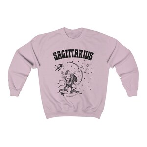 May include: Pale pink sweatshirt with a black Sagittarius design. The graphic shows an archer with a bow and arrow, constellations, and the word "SAGITTARIUS". The design is a vintage style.