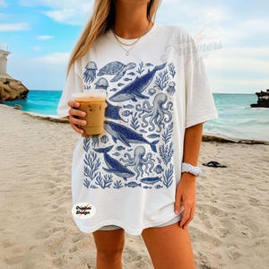 May include: White t-shirt with a blue marine life design, including whales, octopuses, and fish. The shirt has a loose fit and short sleeves. The words "Original Design" are printed on the bottom left of the shirt.