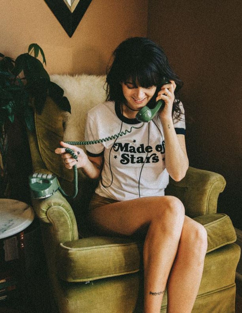 May include: A woman sits in a green armchair and talks on a vintage green rotary phone. She is wearing a white t-shirt with the text "Made of Stars" printed on it.