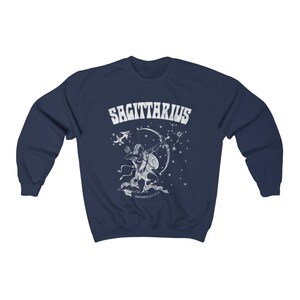 May include: Navy blue sweatshirt with a white Sagittarius zodiac design. The graphic depicts an archer with a bow and arrow, the word "SAGITTARIUS", and star constellations. A comfortable garment for those interested in astrology.