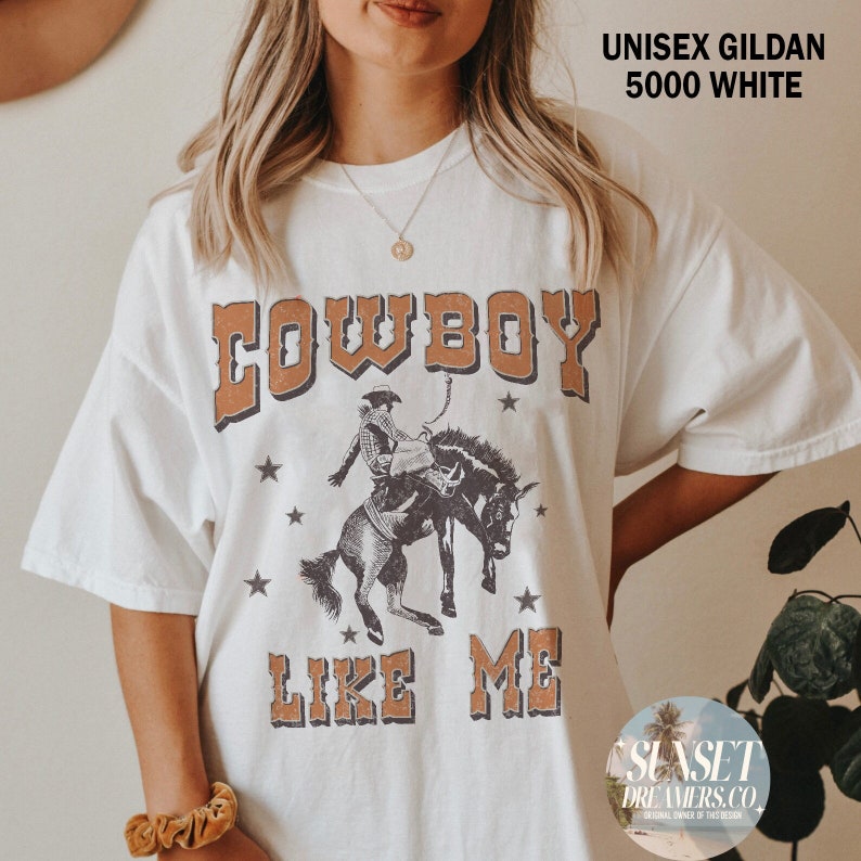 May include: White Gildan 5000 t-shirt with a vintage-style graphic. The design features the word "COWBOY" in brown block letters above an illustration of a cowboy riding a horse, with the words "LIKE ME" below. The shirt is unisex.