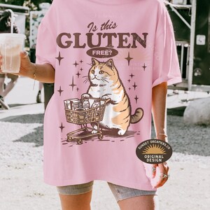 May include: Pink t-shirt with the text "Is this Gluten Free?" and a cartoon cat pushing a shopping cart filled with groceries. The cat is orange and white. The shirt also has star graphics.