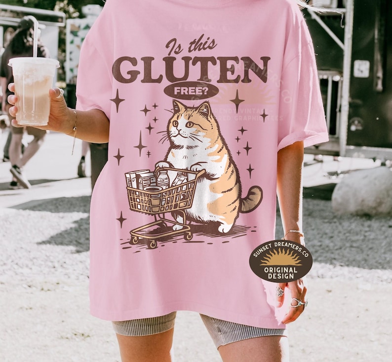 May include: Pink t-shirt with the text "Is this Gluten Free?" and a cartoon cat pushing a shopping cart filled with groceries. The cat is orange and white. The shirt also has star graphics.