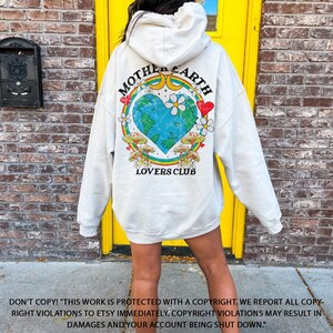 May include: A white hoodie with a graphic print of a heart-shaped Earth with a rainbow, mushrooms, and the text "Mother Earth Lovers Club".