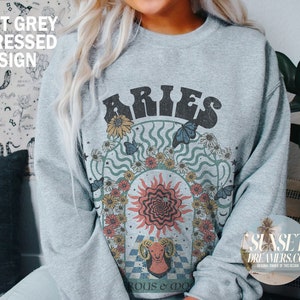 Puede incluir: Una sudadera gris con un dise&ntilde;o desgastado que presenta el signo zodiacal Aries rodeado de flores y mariposas. El texto "Aries" est&aacute; en una fuente retro.