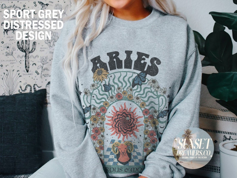 Puede incluir: Una sudadera gris con un dise&ntilde;o desgastado que presenta el signo zodiacal Aries rodeado de flores y mariposas. El texto "Aries" est&aacute; en una fuente retro.