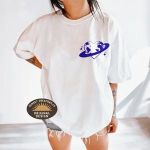 May include: White t-shirt with a blue graphic of a heart-shaped planet with stars. The text "Sunset Dreamers Co. Original Design" is on a brown oval sticker.
