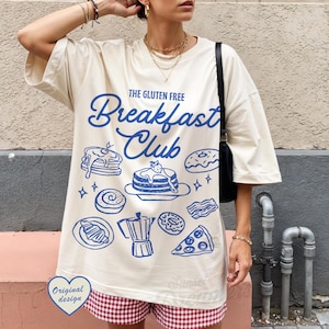 May include: Cream-colored t-shirt with a blue graphic design featuring breakfast foods and the text "The Gluten Free Breakfast Club." The design includes pancakes, donuts, and a coffee pot. The shirt has short sleeves.