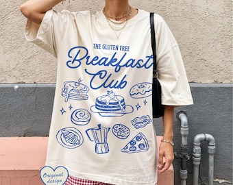 May include: Cream-colored t-shirt with a blue graphic design featuring breakfast foods and the text "The Gluten Free Breakfast Club." The design includes pancakes, donuts, and a coffee pot. The shirt has short sleeves.