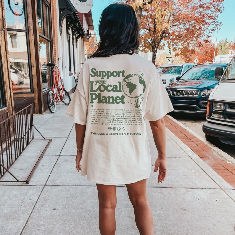 Peut inclure: Une personne portant un t-shirt cr&egrave;me avec un graphisme vert qui dit "Support your Local Planet" et une illustration verte de la Terre. Le texte sous le graphisme dit "Planet Earth is one of the most unique planets in our solar system. It has this wild mix of awesome adventure events and incredible natural beauty. We are all connected to this planet, although the Earth provides nearly endless possibilities, we need to keep things covered in beautiful rock. As many people know, we are all connected to this planet. We can walk, bike, or even walk short distances, yet so many people drive. Take only what you need and enjoy what we have. Embrace a sustainable future."
