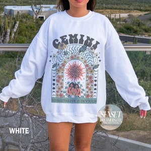 Pode incluir: Camisola branca com um design gr&aacute;fico retro que apresenta o signo do zod&iacute;aco G&eacute;meos, um sol, flores e o texto "Imaginative & Devious". O gr&aacute;fico est&aacute; em tons de azul, verde, amarelo, rosa e laranja. O texto "SUNSET DREAMERS.CO" est&aacute; impresso no canto inferior direito.