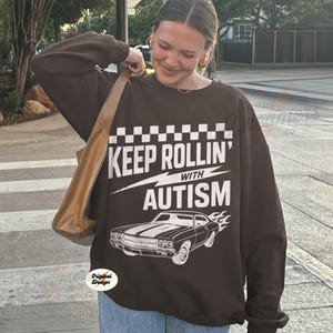 May include: A brown sweatshirt with the text "KEEP ROLLIN' WITH AUTISM" in white, along with a graphic of a classic car with flames. The sweatshirt has a checkered pattern at the top and is a casual style.